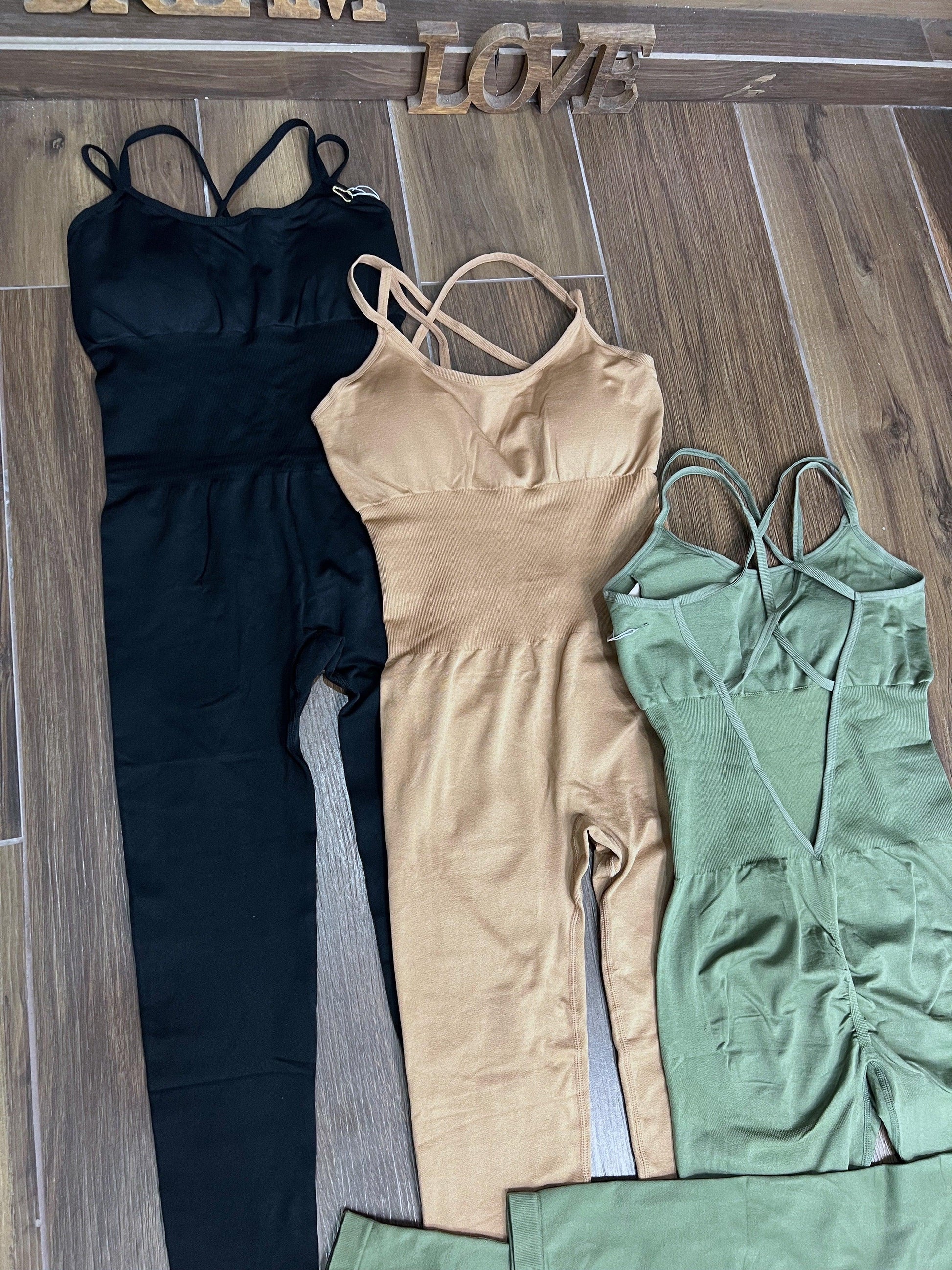 Three SPORT 03 sleeveless fitted jumpsuits in black, beige, and green laid on wooden floor.
