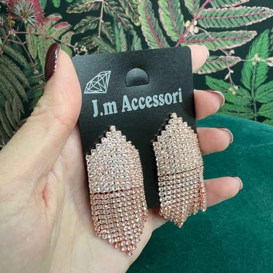 Hand holding rose gold sparkly chandelier earrings on black J.m Accessori card with green floral background