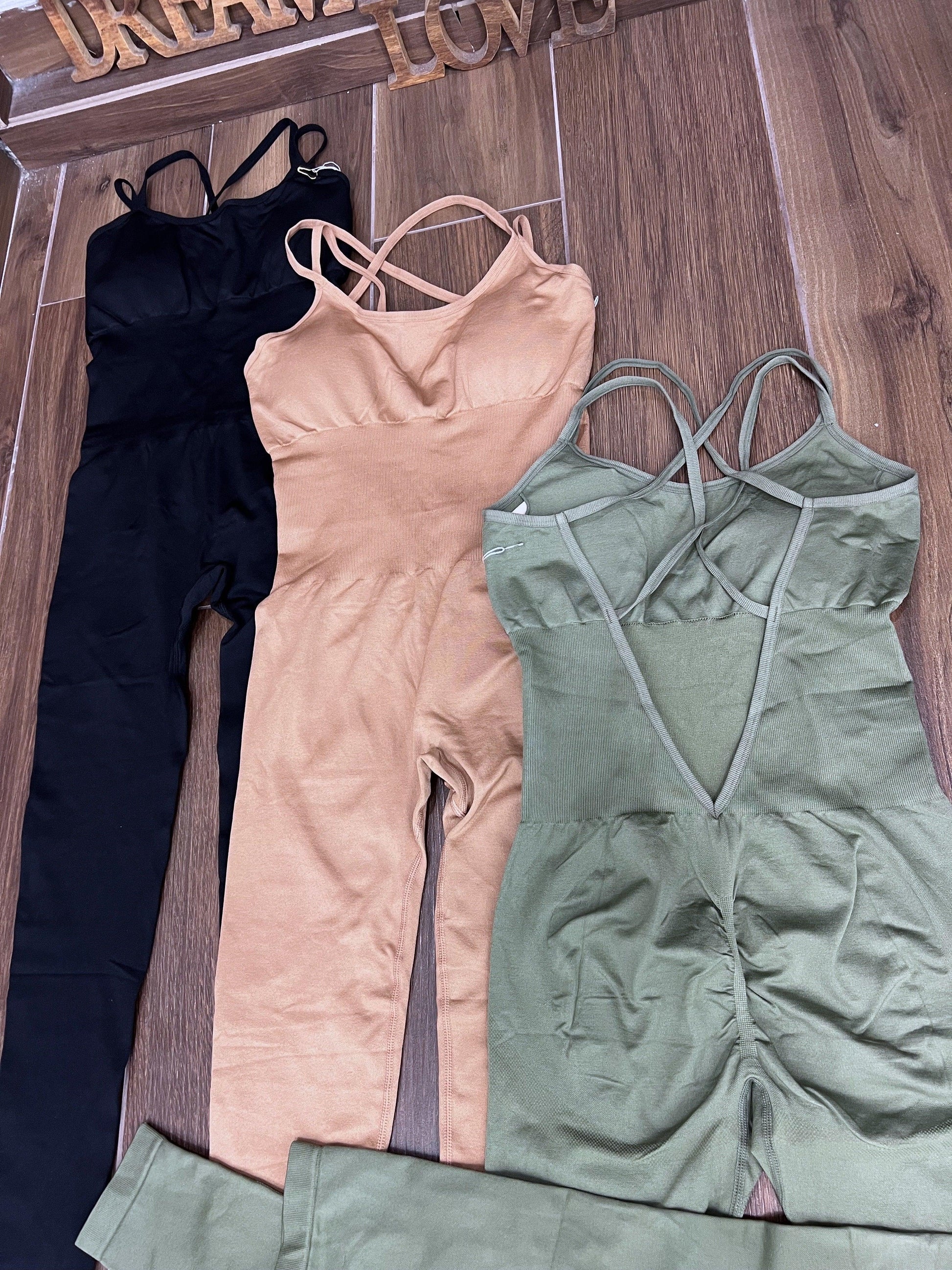 Three SPORT 03 sleeveless workout jumpsuits in black, beige, and olive green laid flat on wooden floor.