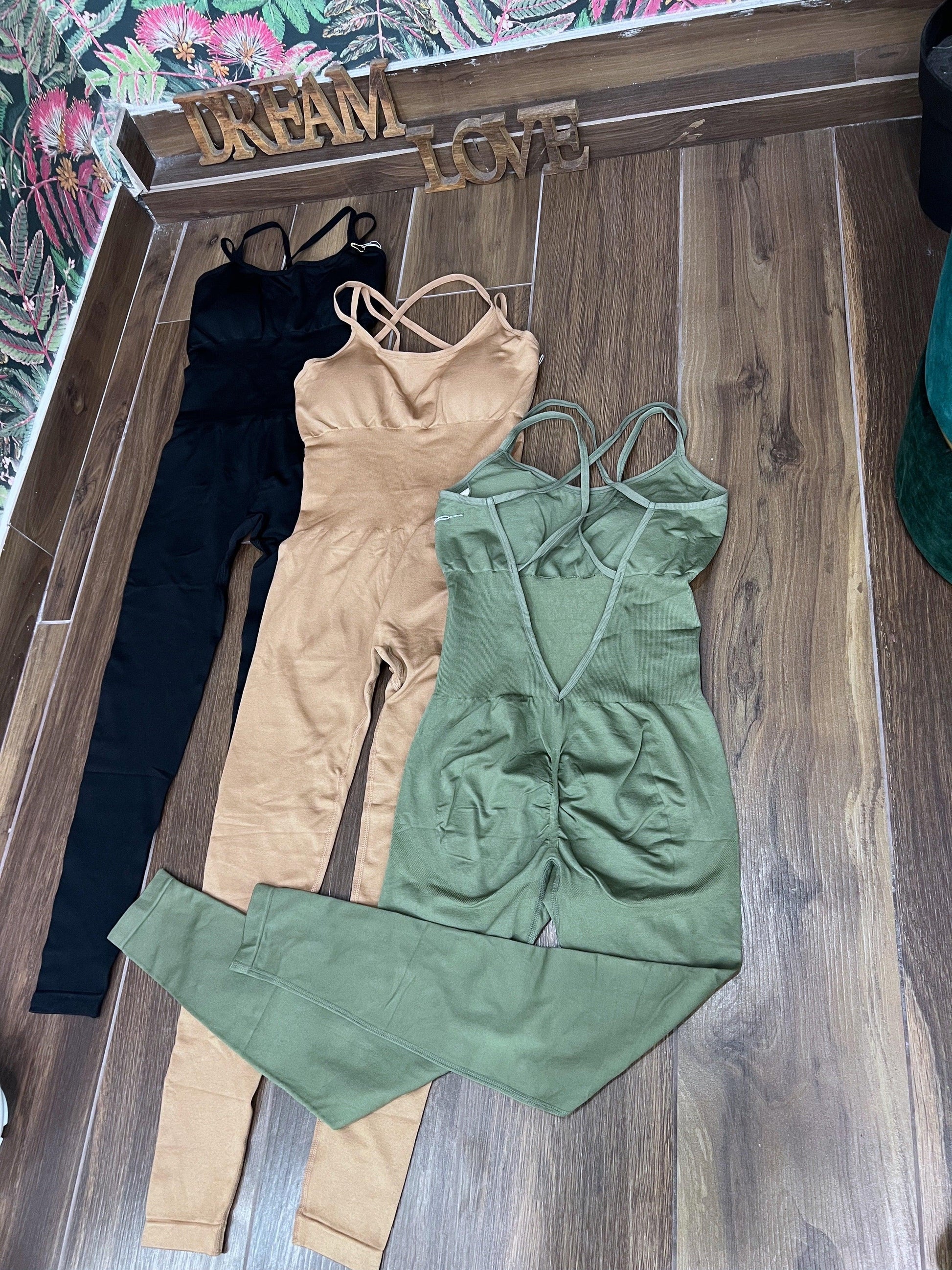 Three SPORT 03 women's one-piece athletic jumpsuits in black, beige, and olive green laid on wooden floor