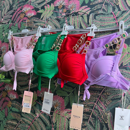 Four colorful bikini tops in pink, green, red, and purple with jeweled straps hanging against floral wallpaper background