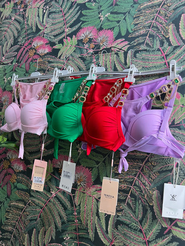 Four colorful bikini tops in pink, green, red, and purple with jeweled straps hanging against floral wallpaper background