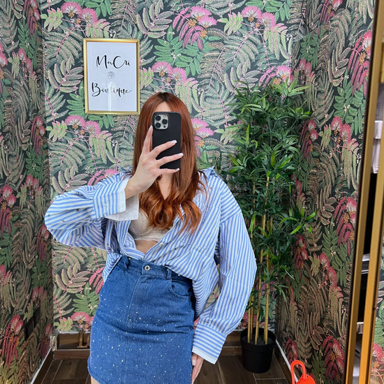 Woman wearing blue striped oversized shirt and denim mini skirt taking a mirror selfie in floral wallpapered room