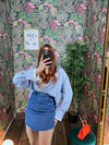 Woman wearing blue striped oversized shirt and denim mini skirt taking a mirror selfie in floral wallpapered room