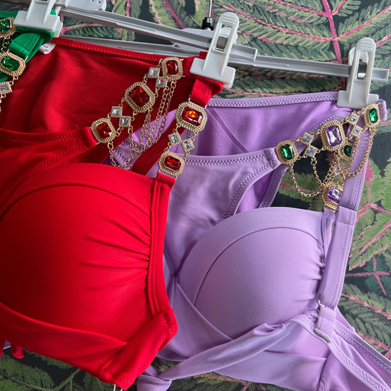 Red and lavender bikini tops with jeweled straps hanging on white hangers against a leafy background