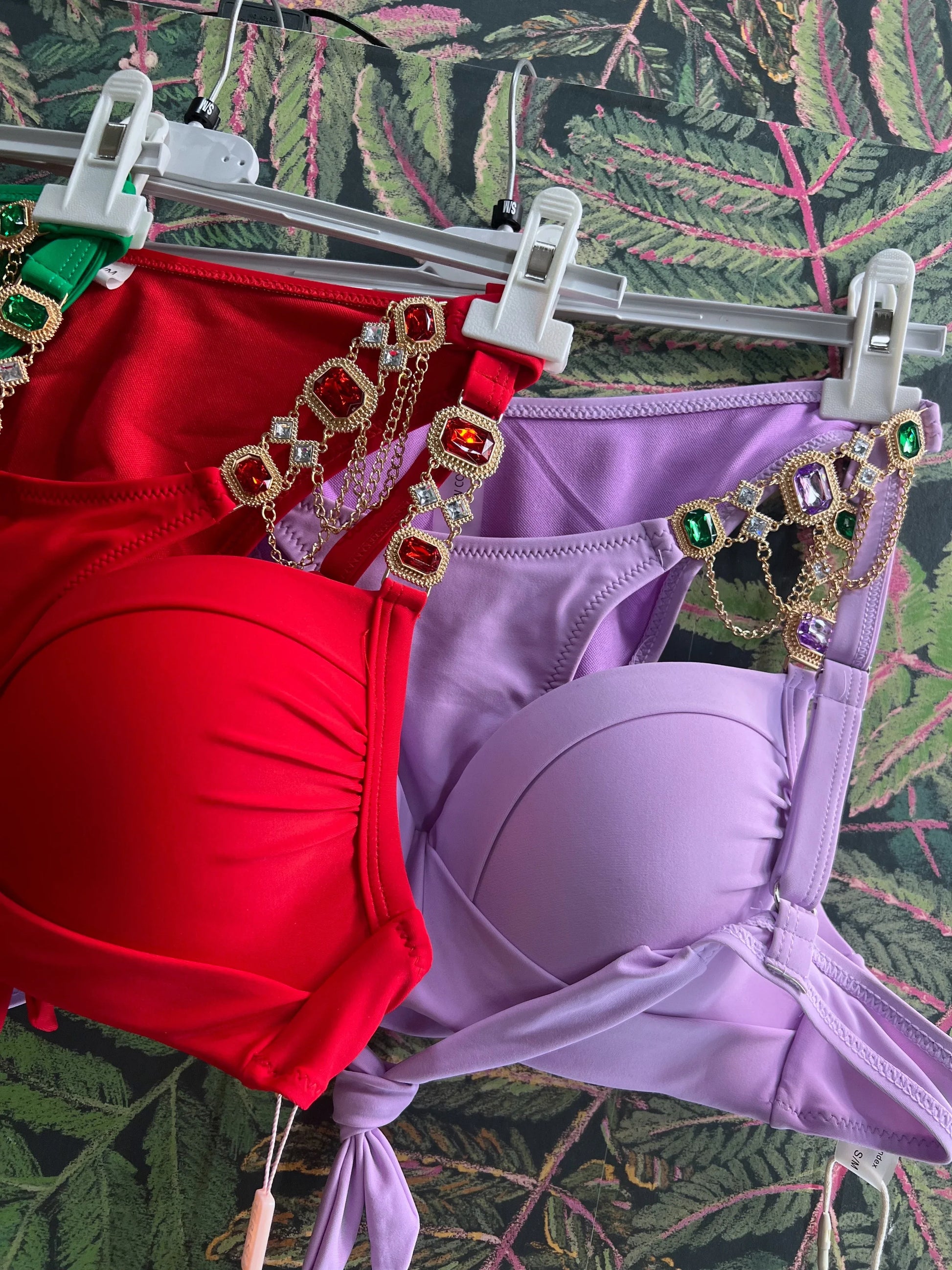 Red and lavender bikini tops with jeweled straps hanging on white hangers against a leafy background