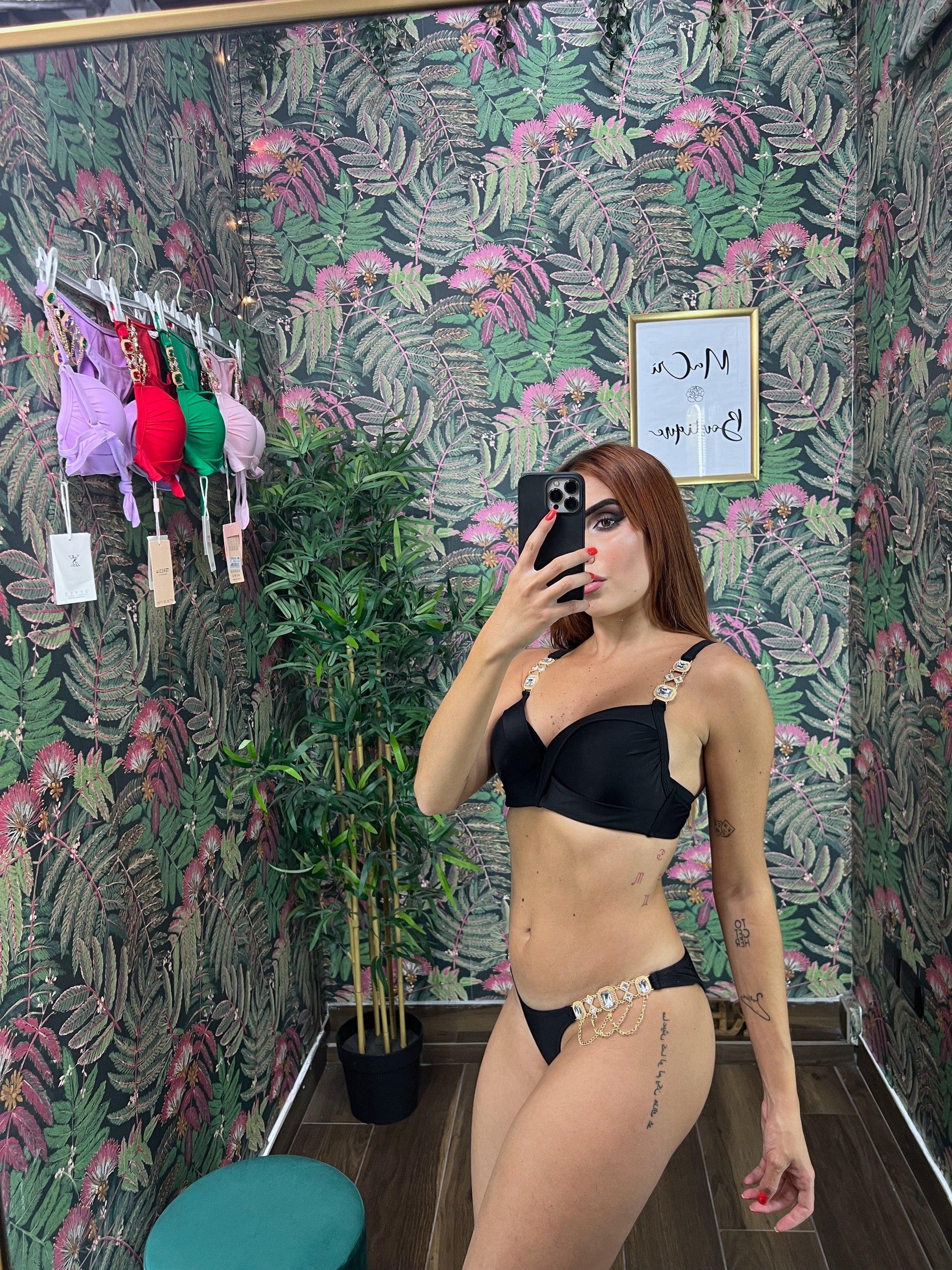 Woman taking mirror selfie wearing black Bikini India in a room with leaf-patterned wallpaper and hanging bikinis.