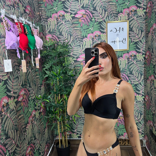 Woman taking mirror selfie wearing black bikini with gold chain details against floral wallpaper background