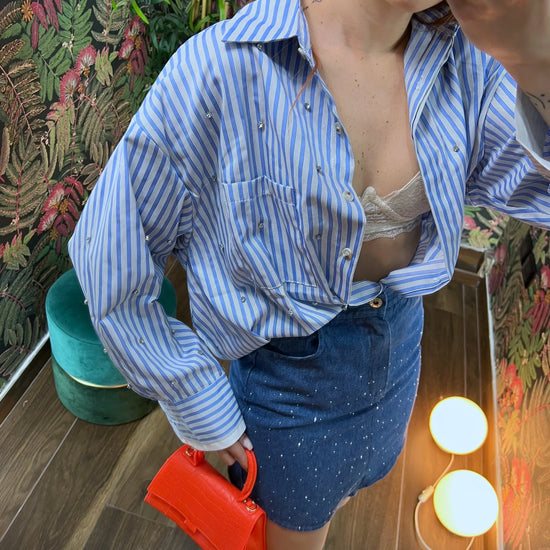 Woman wearing a blue striped shirt, denim skirt, and holding a small red handbag in a floral room setting