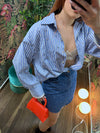 Woman wearing a blue striped shirt, denim skirt, and holding a small red handbag in a floral room setting