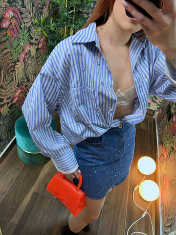 Woman wearing a blue striped shirt, denim skirt, and holding a small red handbag in a floral room setting