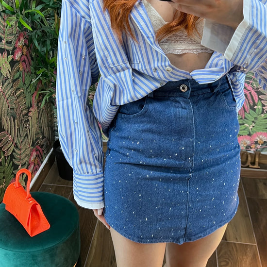 woman wearing blue striped shirt and embellished denim mini skirt standing indoors taking a selfie