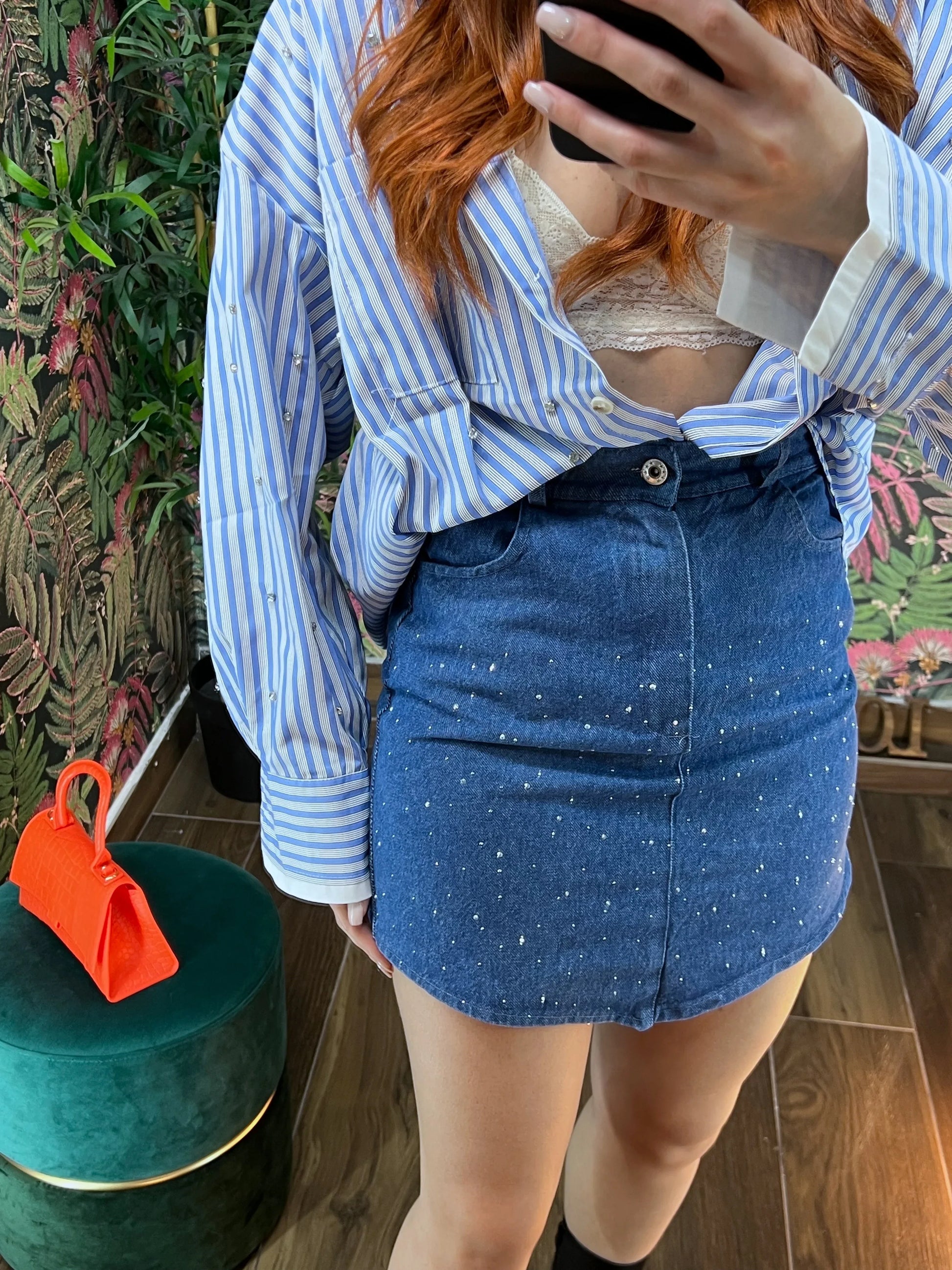 woman wearing blue striped shirt and embellished denim mini skirt standing indoors taking a selfie