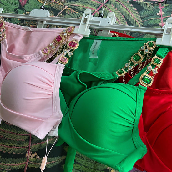 Three colorful bikini tops with jeweled straps hanging on white hangers against a floral background
