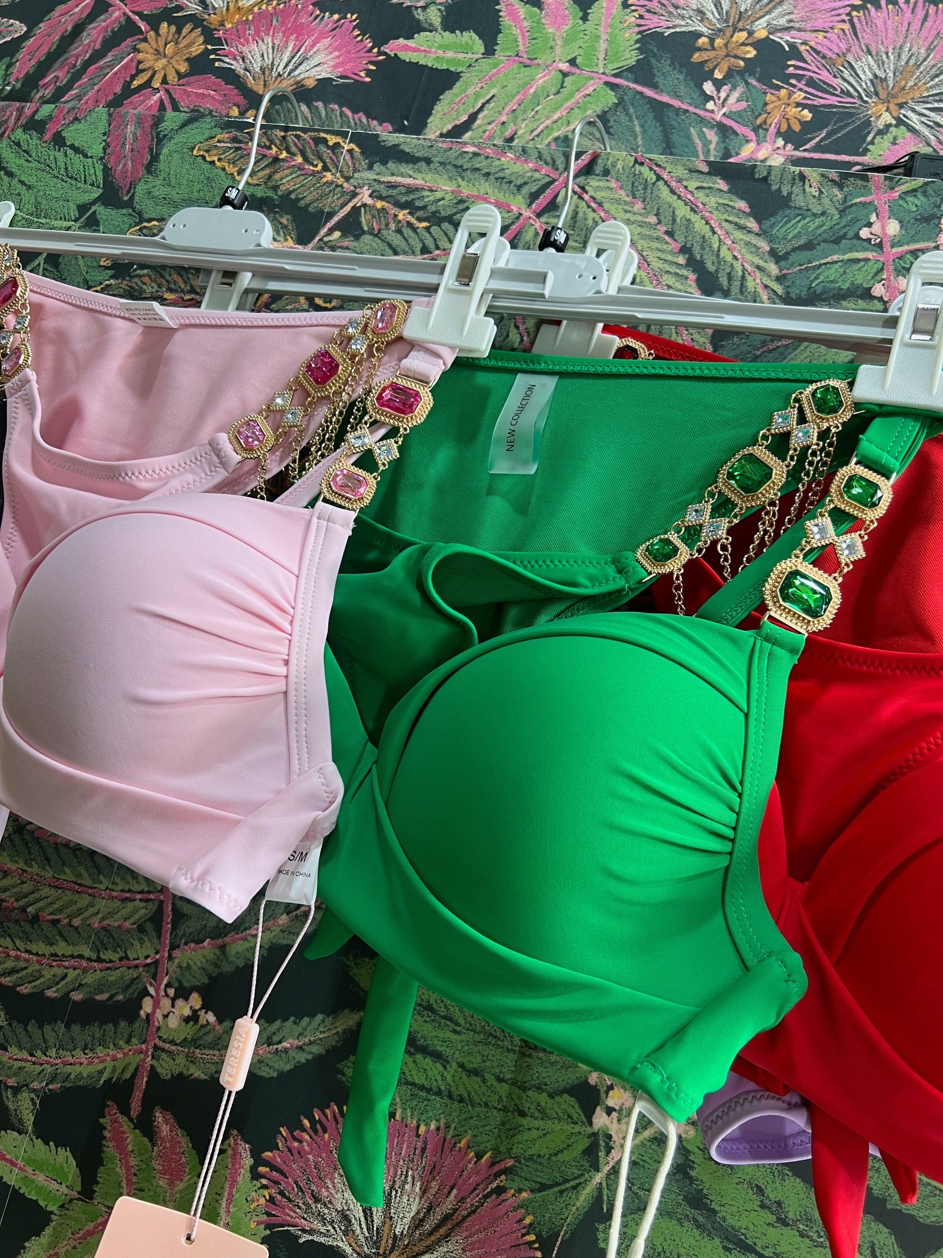 Three colorful bikini tops with jeweled straps hanging on white hangers against a floral background
