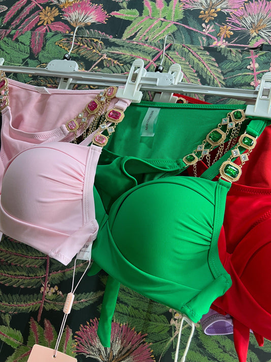 Three colorful bikini tops with jeweled straps hanging on white hangers against a floral background