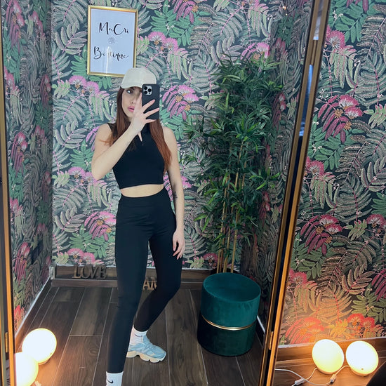 Woman in black fitness crop top and leggings taking a mirror selfie in a tropical-themed room with sneakers and cap
