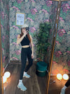 Woman in black fitness crop top and leggings taking a mirror selfie in a tropical-themed room with sneakers and cap