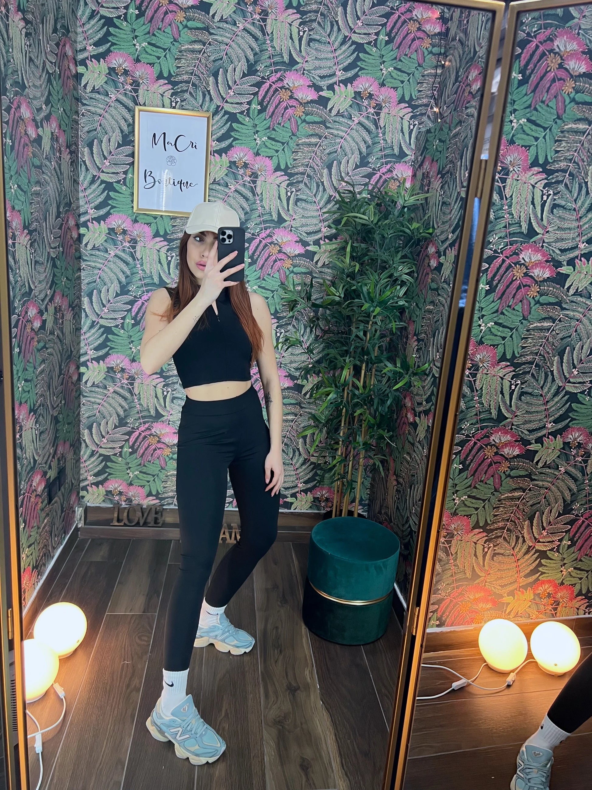 Woman in black fitness crop top and leggings taking a mirror selfie in a tropical-themed room with sneakers and cap