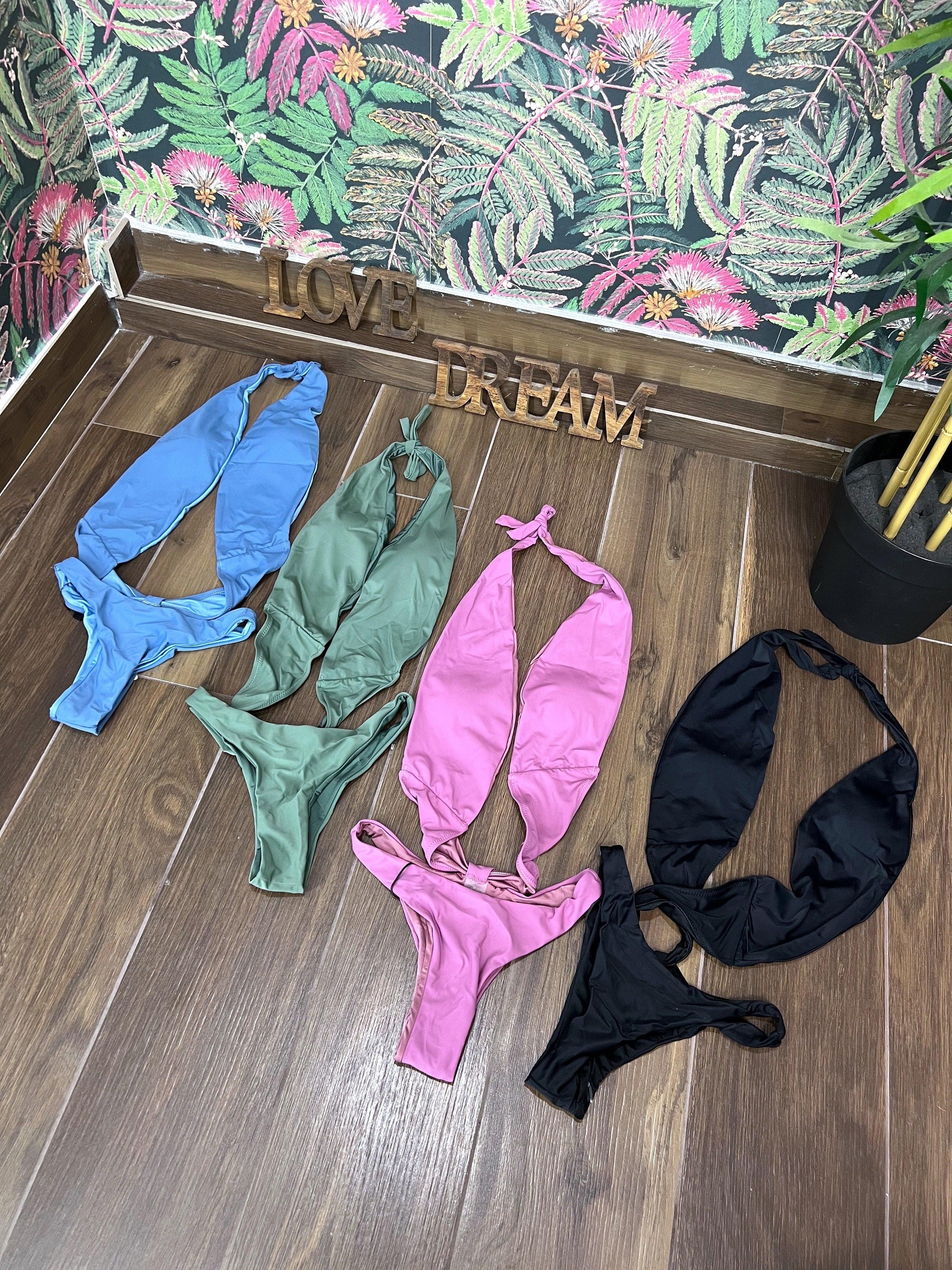 Four trikini swimsuits in blue, green, violet, and black laid out on a wooden floor with floral wallpaper background.