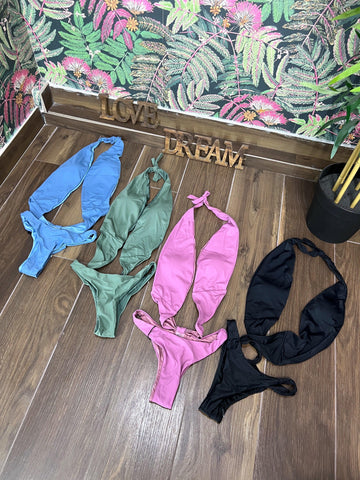 Four trikini swimsuits in blue, green, violet, and black laid out on a wooden floor with floral wallpaper background.