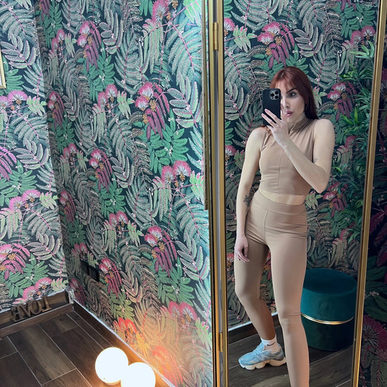 Woman wearing beige fitness outfit posing in front of a mirror with tropical patterned wallpaper background