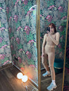 Woman wearing beige fitness outfit posing in front of a mirror with tropical patterned wallpaper background