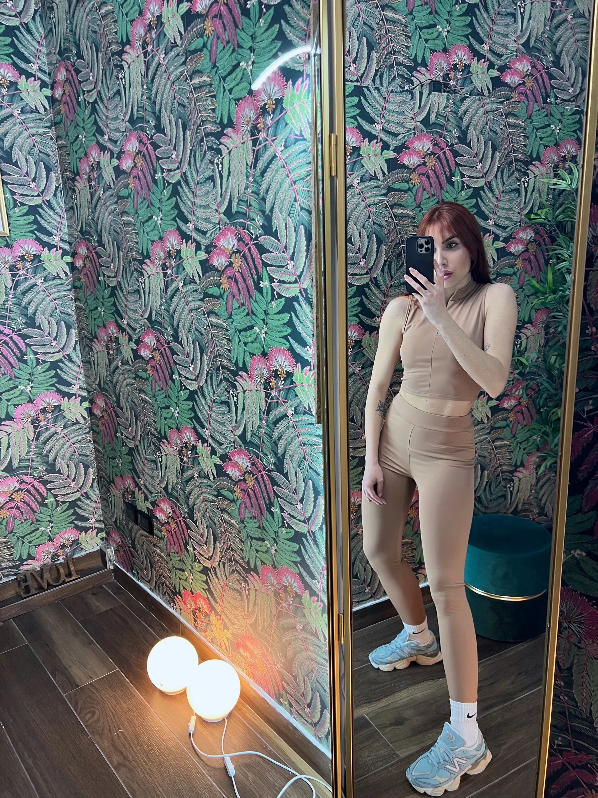 Woman wearing beige fitness outfit posing in front of a mirror with tropical patterned wallpaper background