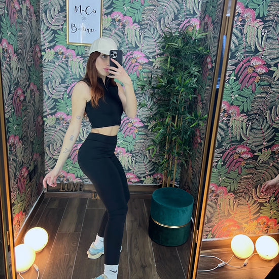 Woman wearing black fitness outfit and white cap taking mirror selfie in a tropical-themed room with plants and decor