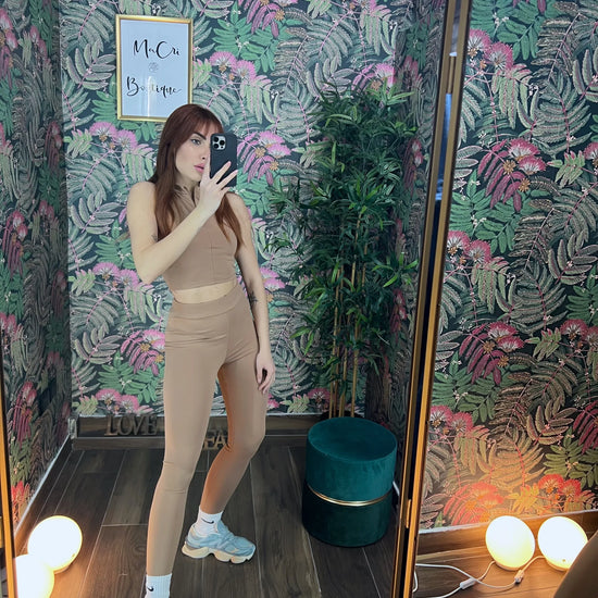 Woman taking mirror selfie wearing tan coordinated fitness outfit with light blue sneakers in floral wallpaper room