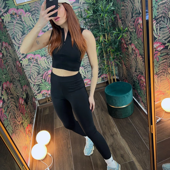 Woman wearing black fitness crop top and leggings taking mirror selfie in floral wallpapered room