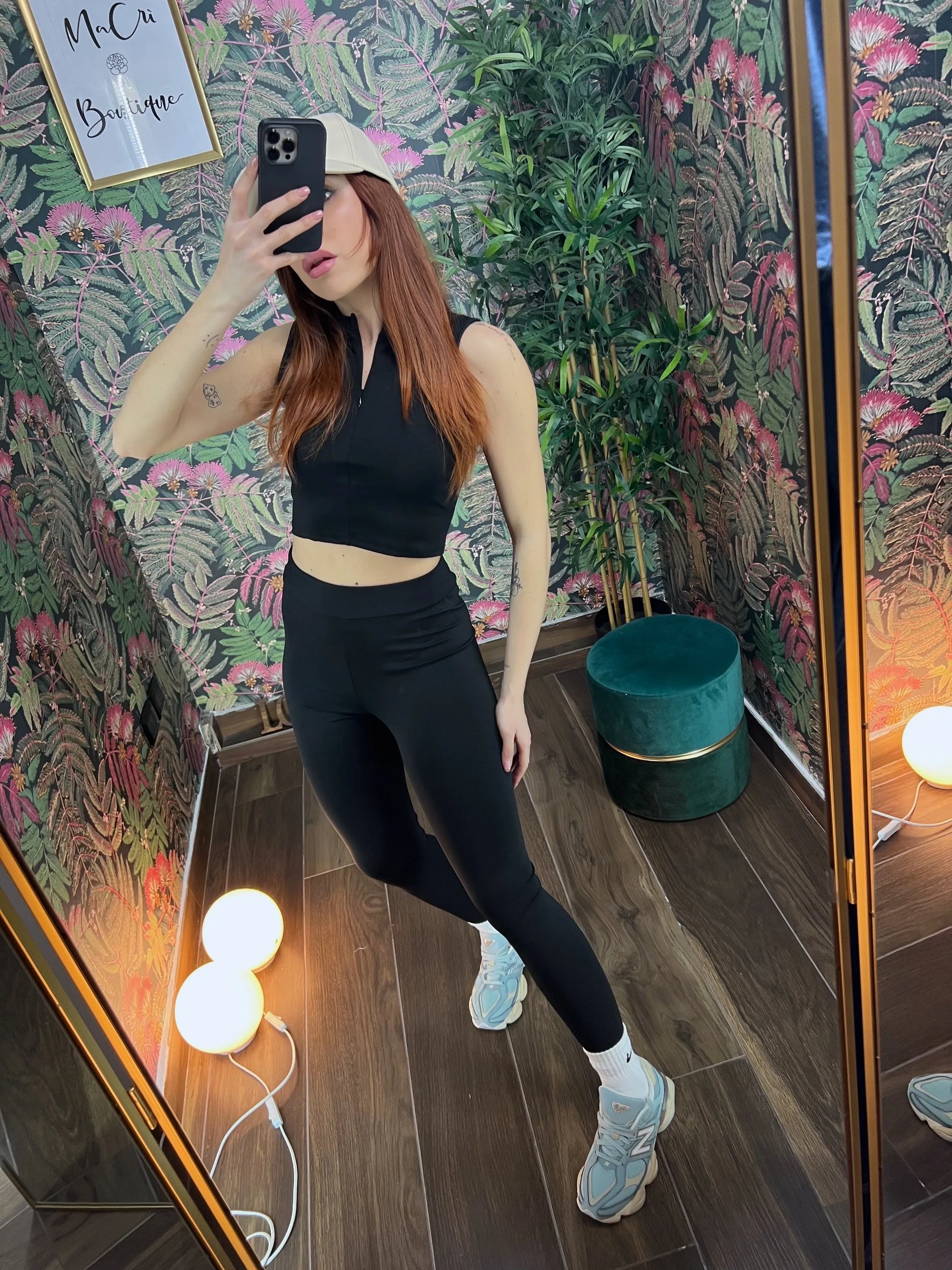 Woman wearing black fitness crop top and leggings taking mirror selfie in floral wallpapered room