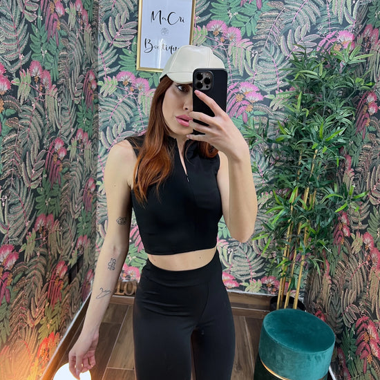Woman in black sleeveless fitness crop top and leggings taking a mirror selfie wearing a white cap in a floral room