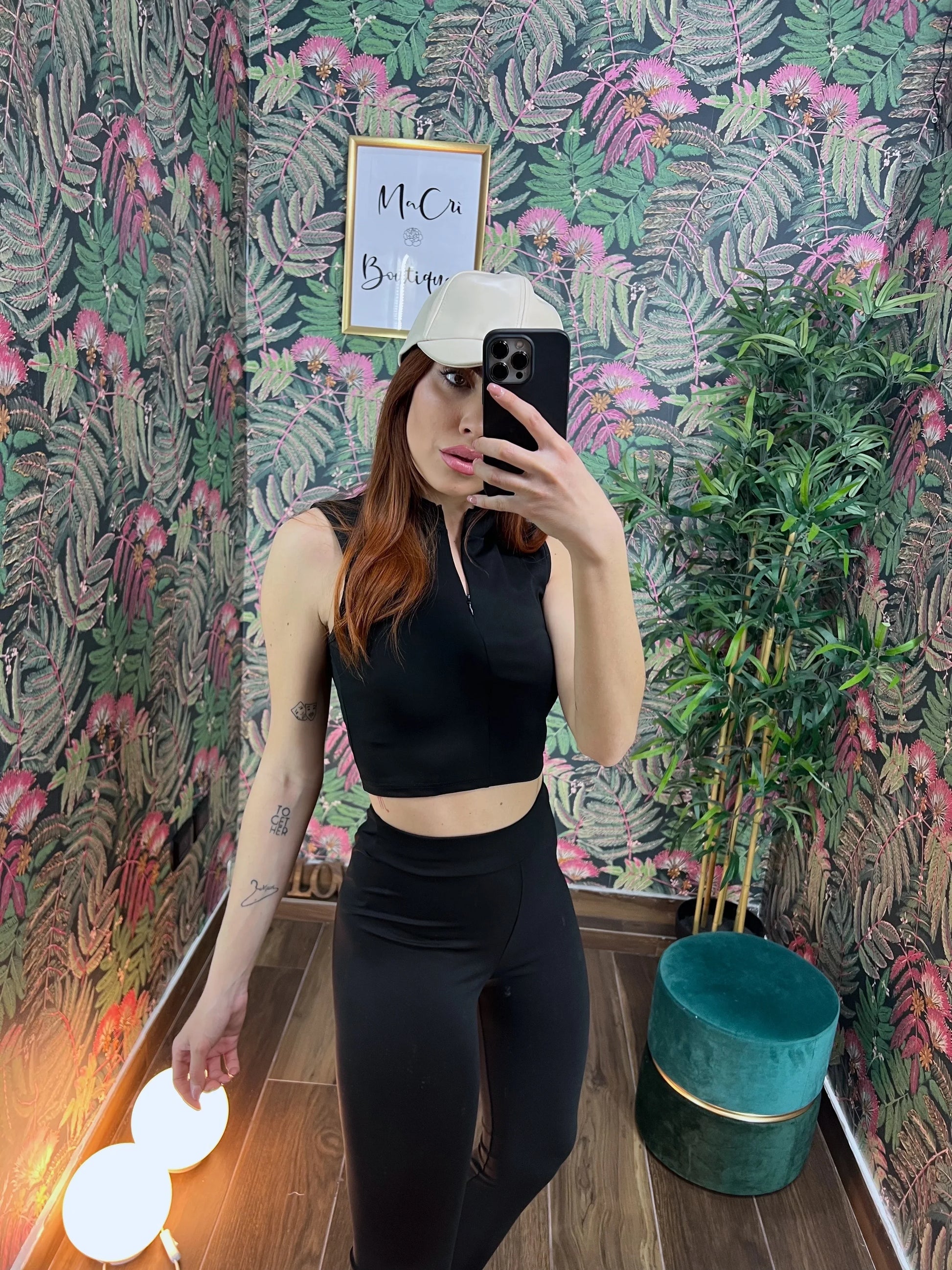 Woman in black sleeveless fitness crop top and leggings taking a mirror selfie wearing a white cap in a floral room