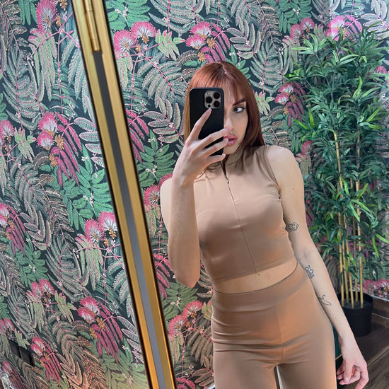Woman taking a mirror selfie wearing a beige sleeveless crop top and high-waisted leggings fitness outfit indoors