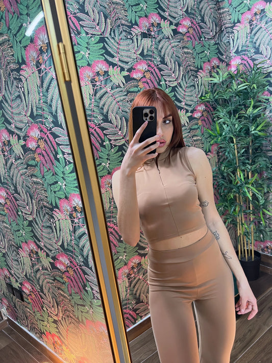 Woman taking a mirror selfie wearing a beige sleeveless crop top and high-waisted leggings fitness outfit indoors
